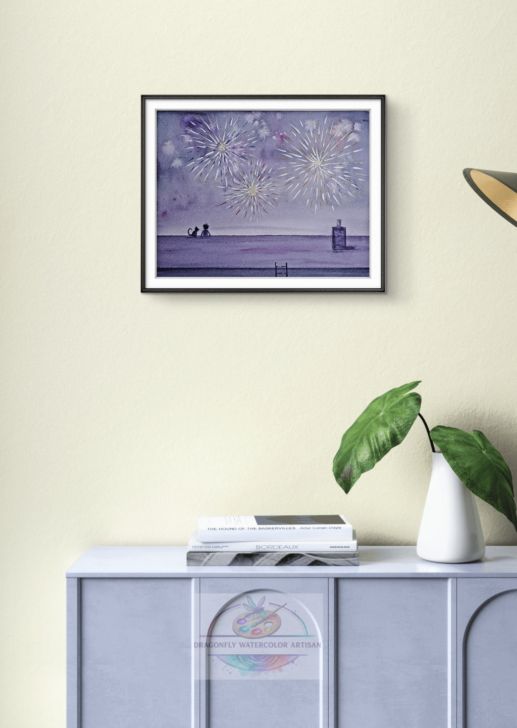 Original watercolor painting titled "Fireworks & Friends" in a thin black frame with white mat, hanging on a light-colored wall above a console table. Features golden fireworks exploding across a deep indigo sky with sweet silhouettes of a child and cat watching from a rooftop.