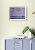 Original watercolor painting titled "Fireworks & Friends" in a thin black frame with white mat, hanging on a light-colored wall above a console table. Features golden fireworks exploding across a deep indigo sky with sweet silhouettes of a child and cat watching from a rooftop.