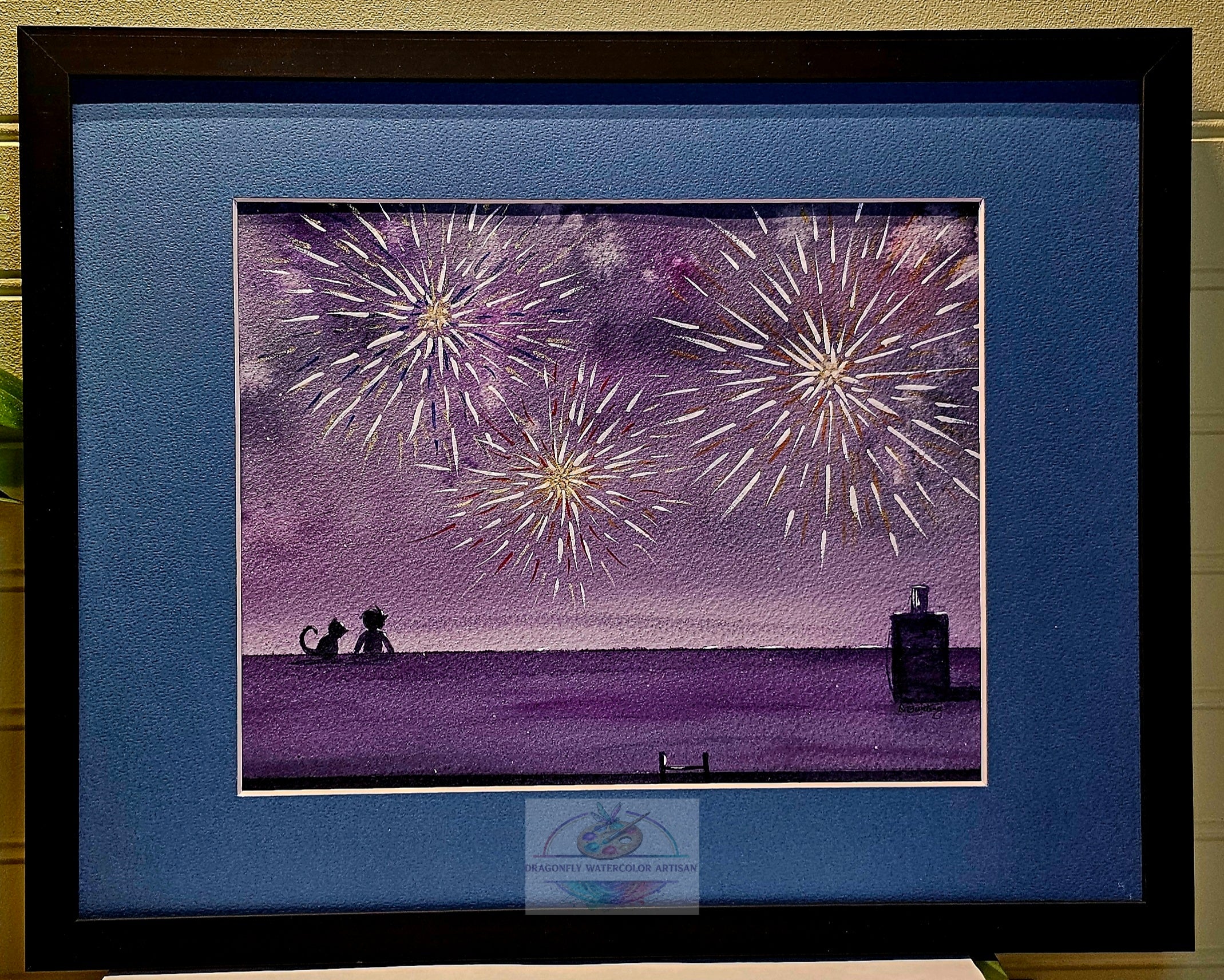 Framed original watercolor "Fireworks & Friends" showing the full piece in a black frame with blue mat. Golden fireworks burst over a purple night sky while a child and cat sit together on a rooftop watching.