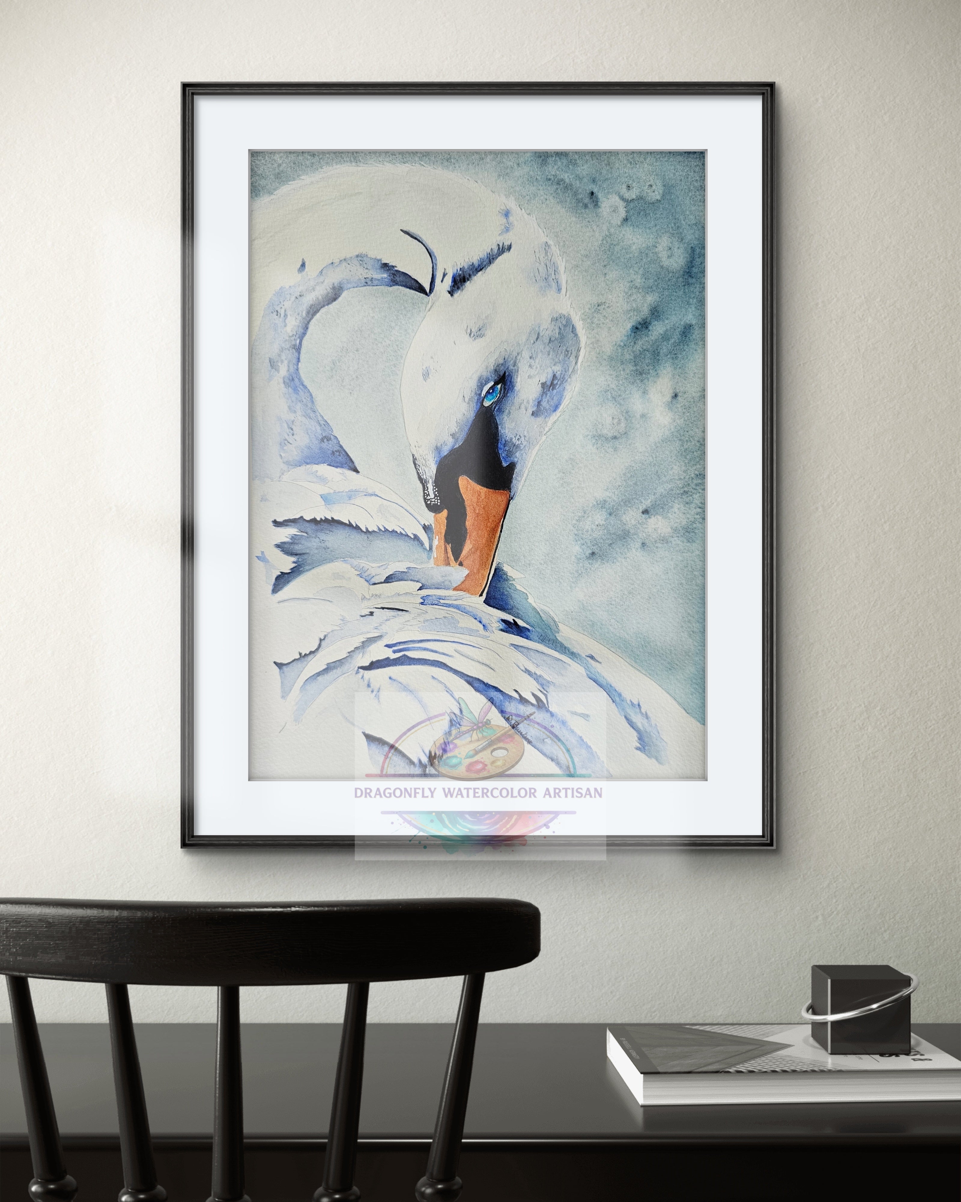 Framed original watercolor swan painting "Grace in Blue" hanging on a light-colored wall above a black chair in a clean, modern interior setting. Shown in a thin black frame with white mat.