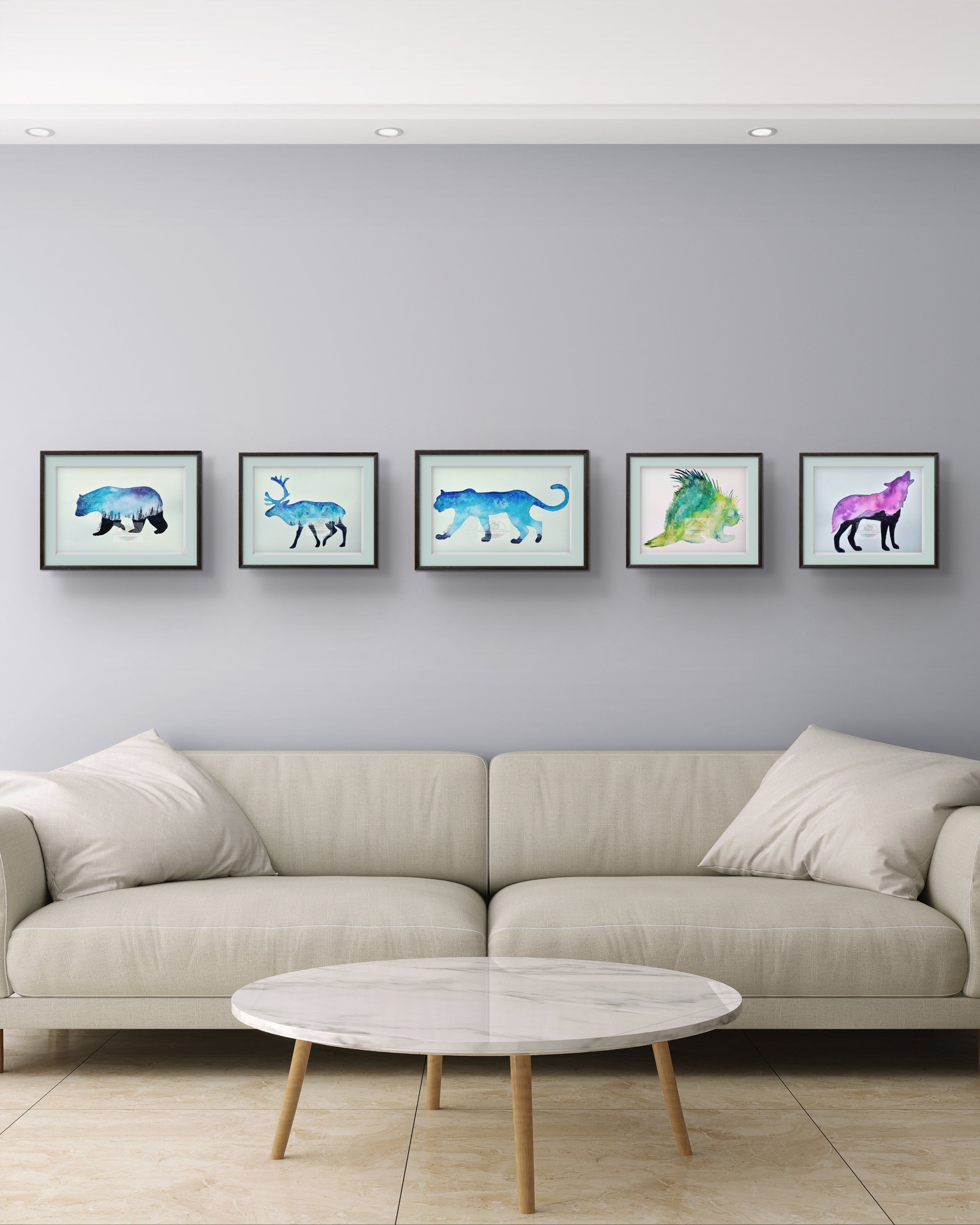 Five framed watercolor animal art prints hung above a white sofa in a modern living room. Blue bear, reindeer, and panther with purple wolf and green porcupine silhouettes. Gallery wall set of wildlife watercolor paintings.