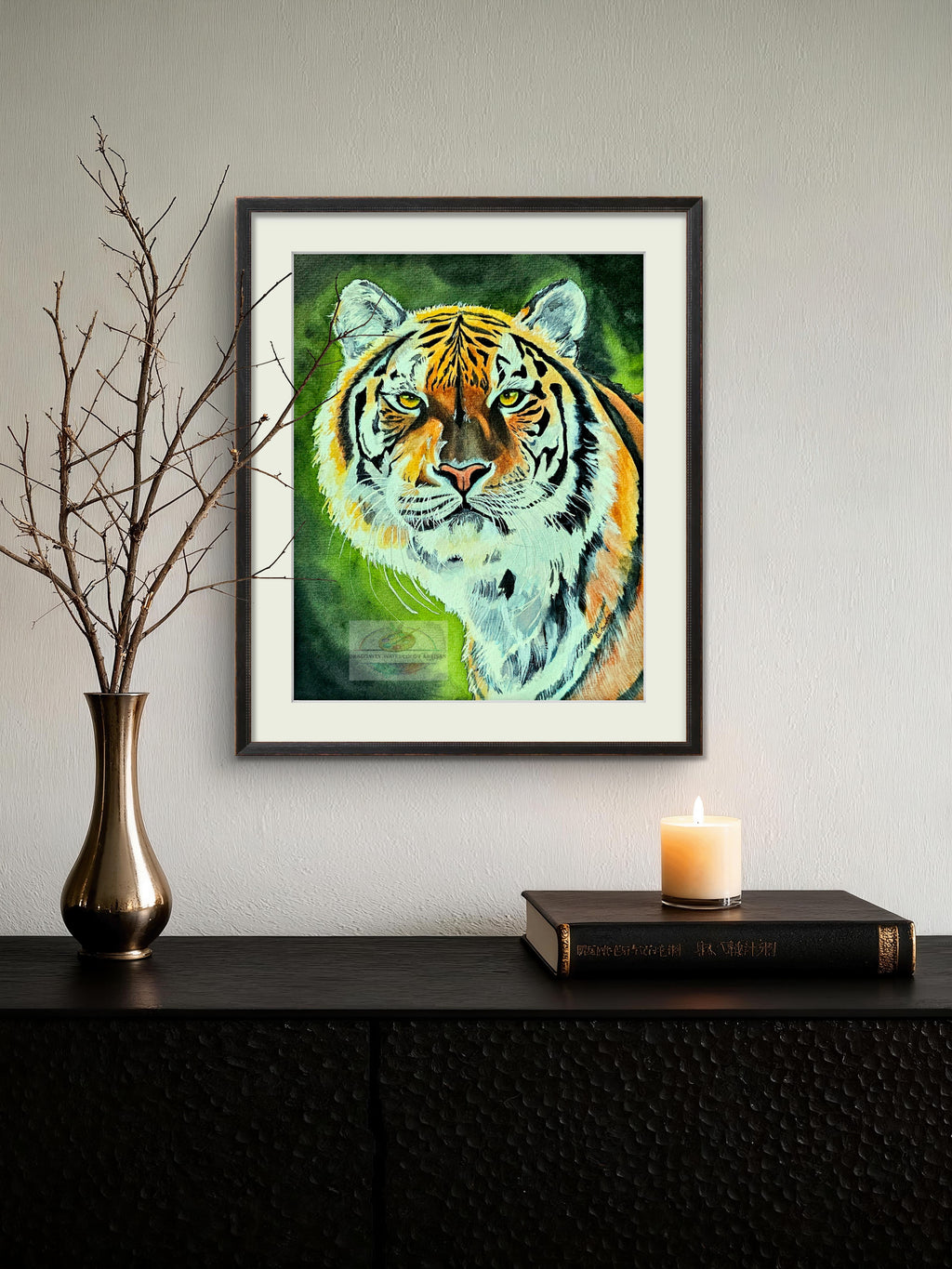 Framed original watercolor tiger portrait hanging on a light wall above a dark console table with a vase and lit candle. Shows the full piece in a dark frame with white mat.